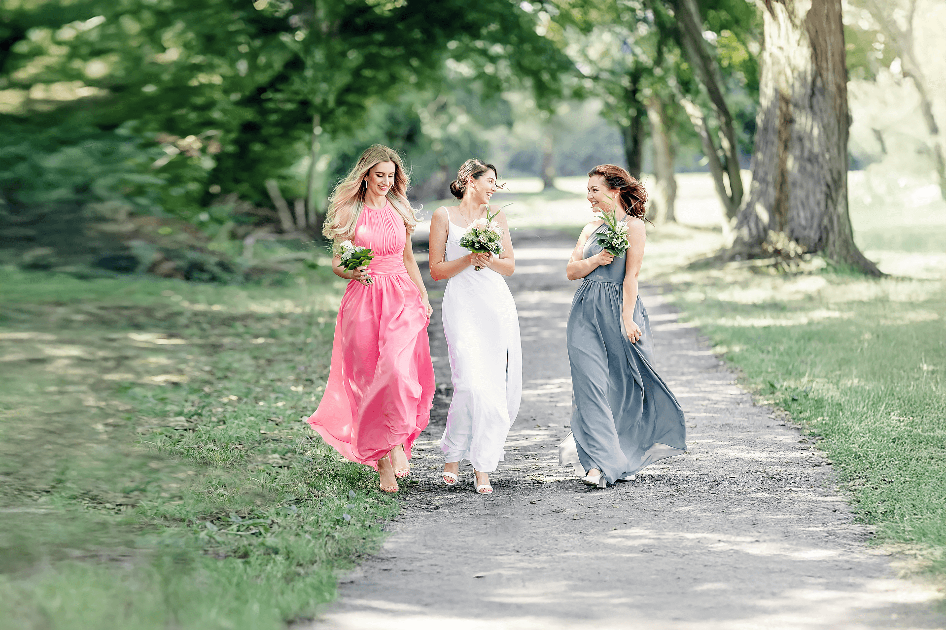 Drei Frauen in eleganten Kleidern - rosa, weiß und blaugrau - gehen gemeinsam auf einem sonnenbeschienenen, von Bäumen gesäumten Weg, jede hält einen kleinen Blumenstrauß und lächelt die andere an.