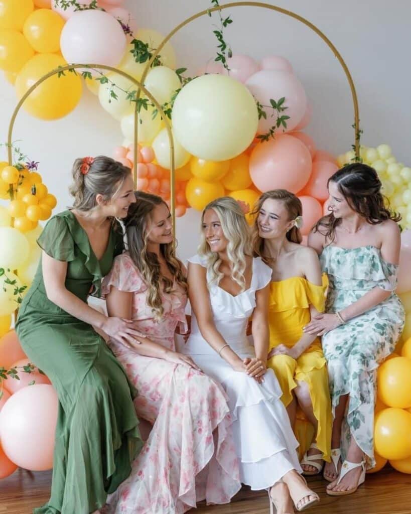 Fünf Frauen in farbenfrohen Brautjungfernkleidern sitzen vor einem pastellfarbenen Ballonbogen mit Grünzeug und lächeln fröhlich zusammen. Eine trägt weiß, die anderen tragen grüne, rosa, gelbe und geblümte Kleider.