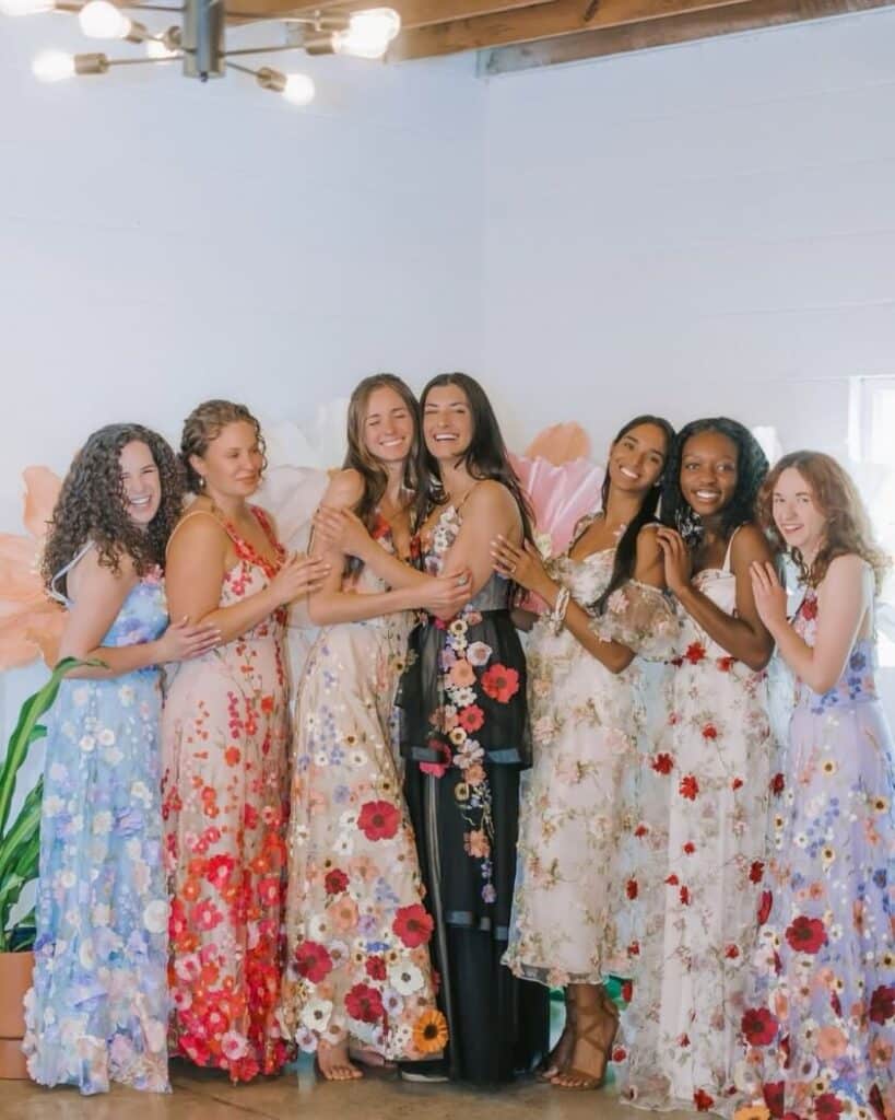 Sieben Frauen stehen in einem Innenraum zusammen, lächeln und umarmen sich in leuchtenden Brautjungfernkleidern. Ihre langen, farbenfrohen, blumengemusterten Kleider ergänzen den pastellfarbenen Blumenschmuck an der Wand und schaffen eine fröhliche und feierliche Atmosphäre.