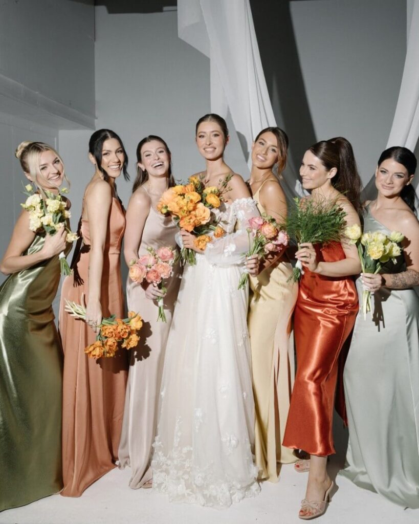 Eine Braut in einem weißen Kleid steht lächelnd mit sechs Brautjungfern in bunten Satin-Brautjungfernkleidern, die jeweils einen Blumenstrauß in der Hand halten. Sie posieren eng beieinander vor einem einfachen, hellen Hintergrund.