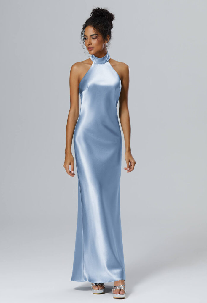 Eine Frau in einem langen, hellblauen Satin-Halfterkleid mit hohem Ausschnitt und offenen Schultern, das an ein elegantes Brautjungfernkleid erinnert, steht in silbernen hochhackigen Sandalen vor einem schlichten hellgrauen Hintergrund.