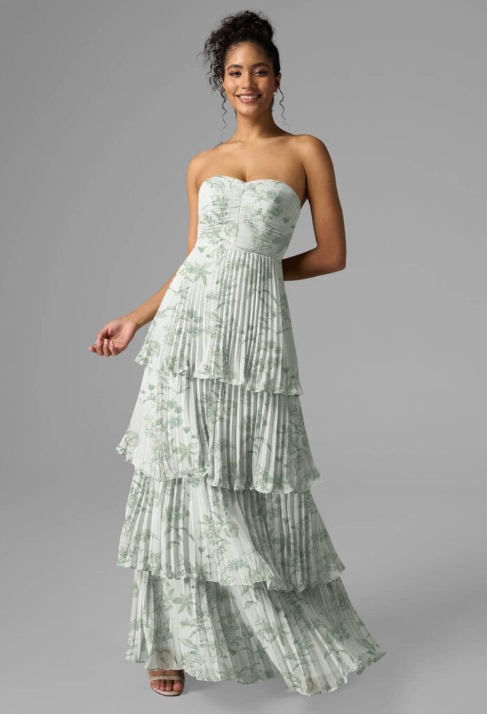 Eine Frau, die ein trägerloses, bodenlanges, gestuftes Kleid mit einem hellgrünen Blumenmuster trägt - perfekt für Brautjungfernkleider -, posiert vor einem grauen Hintergrund und lächelt mit einer erhobenen Hand und der anderen an ihrer Seite.