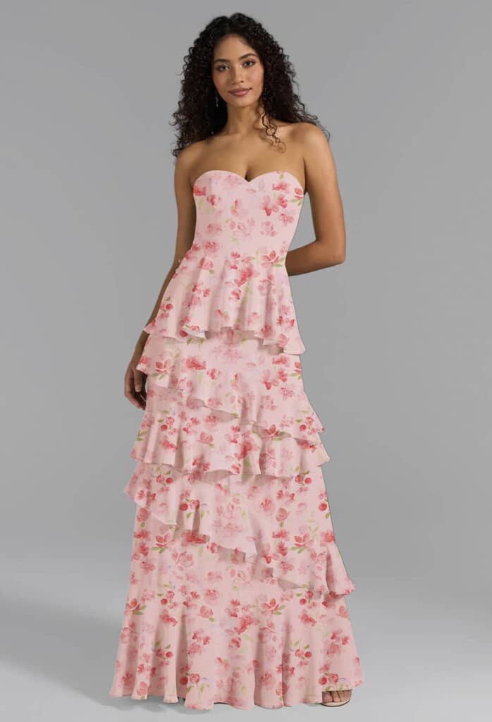 Eine Frau in einem trägerlosen, bodenlangen rosafarbenen Kleid mit herzförmigem Ausschnitt und gestuften Rüschen, verziert mit einem zarten Blumenmuster - perfekt für Brautjungfernkleider - steht vor einem schlichten grauen Hintergrund.