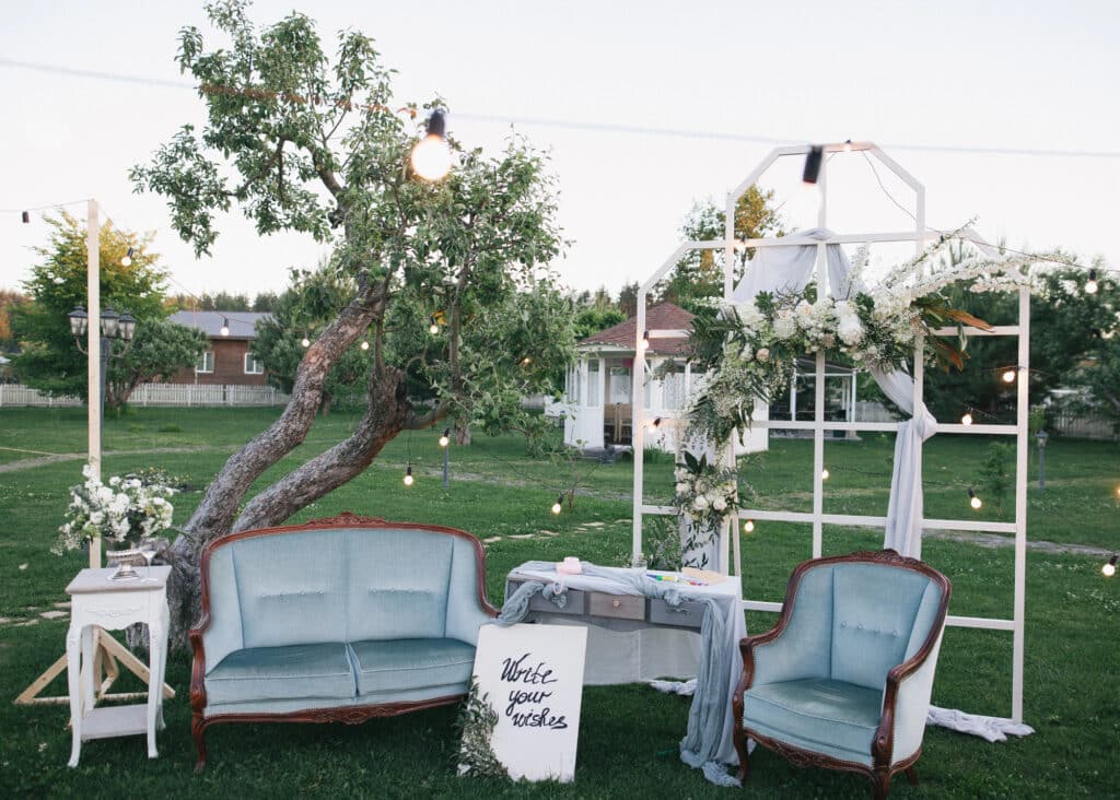 Hochzeit im Freien mit blauen Sofas im Vintage-Stil, einem weißen Tisch mit einem Schild "Schreiben Sie Ihre Hochzeitsglückwünsche", Blumenarrangements und einem dekorativen weißen Bogen, der mit Stoff und Grünzeug unter Lichterketten drapiert ist.