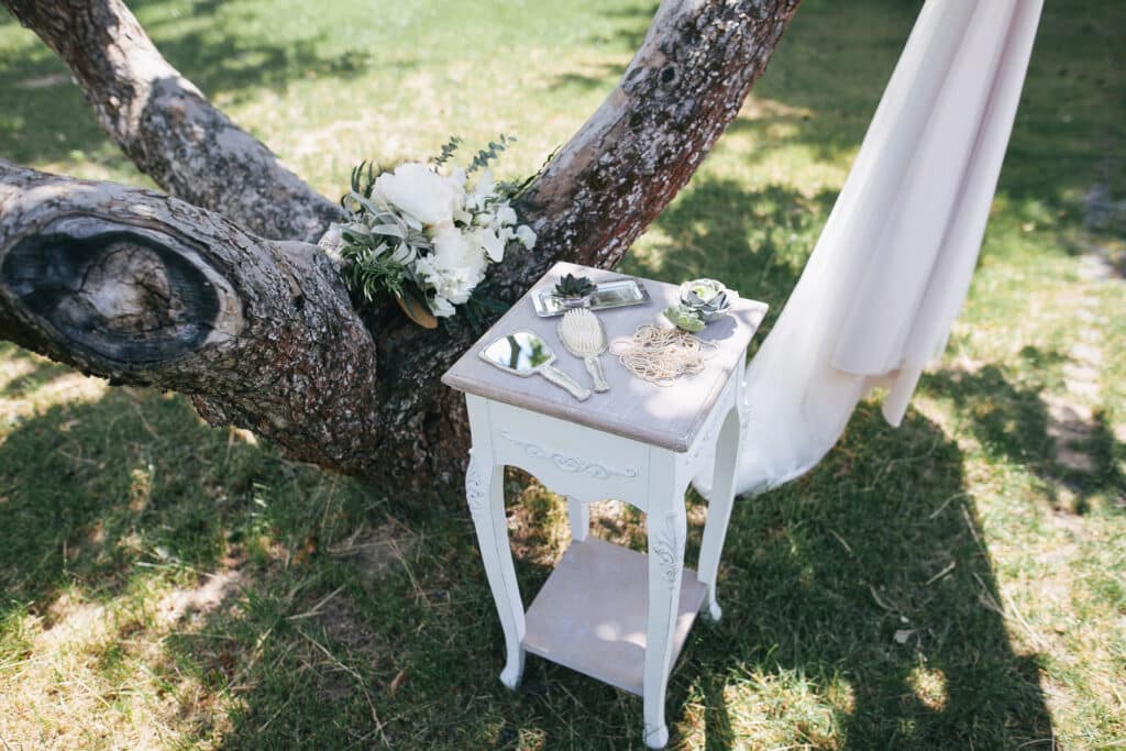 Ein kleiner weißer Tisch mit Dekorationsartikeln und Grünzeug steht neben einem Baum, an dessen Stamm auf einer sonnigen Rasenfläche ein Strauß weißer Blumen und Hochzeitsglückwünsche lehnt.