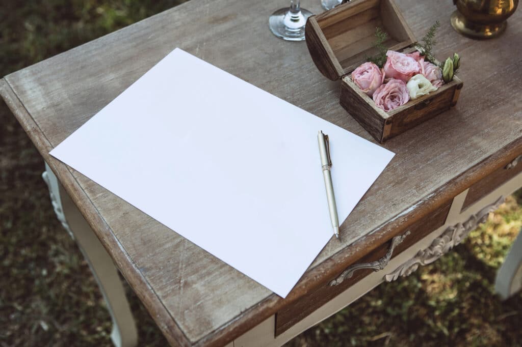 Ein leeres Blatt Papier und ein Stift liegen auf einem hölzernen Schreibtisch neben einer kleinen offenen Schachtel mit rosa und weißen Blumen - eine einladende Umgebung zum Schreiben von Hochzeitsglückwünschen, die in sanftes Sonnenlicht getaucht ist, das die rustikale Atmosphäre im Freien noch verstärkt.