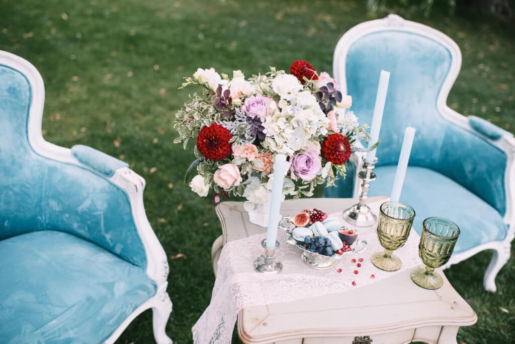 Zwei blaue Samtstühle im Vintage-Stil stehen im Gras, dazwischen ein kleiner Tisch, der mit einem Spitzentuch, einem Blumenstrauß, hohen weißen Kerzen, Obst und zwei grünen Glaspokalen dekoriert ist - perfekt, um Hochzeitsglückwünsche in einer bezaubernden Umgebung zu teilen.