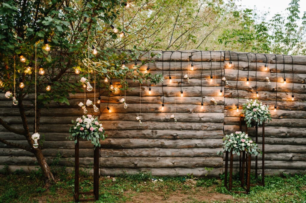 Eine Hochzeitskulisse im Freien mit einer rustikalen Holzblockwand, Lichterketten und hängenden Glühbirnen schafft einen zauberhaften Rahmen für Hochzeitsglückwünsche. Zwei hohe Blumenarrangements mit weißen und rosafarbenen Blumen stehen auf beiden Seiten, umgeben von Grünzeug.
