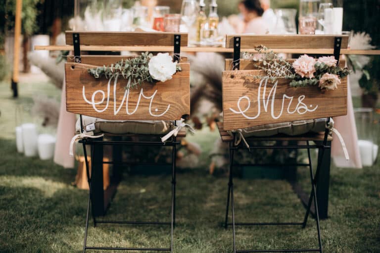 Zwei geschmückte Stühle mit der Aufschrift "Mr." und "Mrs.", an deren Lehnen Blumen und Grünzeug befestigt sind, erwarten das Paar an einem Holztisch mit Getränken - perfekt für gemeinsame Hochzeitsglückwünsche.