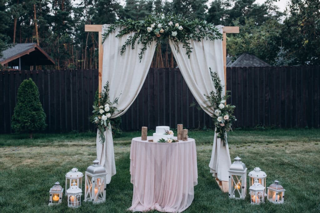 Hochzeit im Freien mit einem hölzernen Bogen, der mit weißem Stoff und Grünzeug drapiert ist, einem runden Tisch mit Torte und weißen Laternen mit Kerzen auf dem Rasen - perfekt, um Hochzeitsglückwünsche unter Bäumen und einem rustikalen Holzzaun zu feiern.