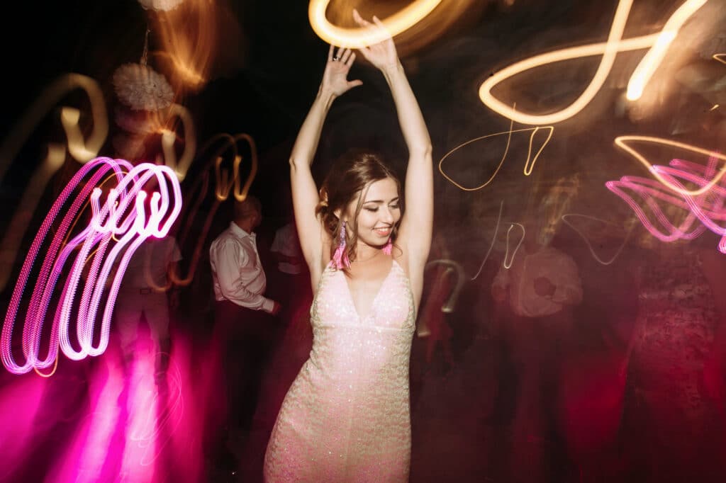 Eine Frau in einem glitzernden Kleid tanzt mit erhobenen Armen, umgeben von leuchtend rosafarbenen und gelben Lichtspuren, während Musik die Luft erfüllt und eine lebhafte und festliche Partyatmosphäre schafft.