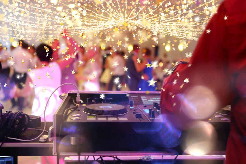Ein DJ im roten Hemd steht an einem Plattenspieler und legt auf einer lebhaften Party Musik auf. Glitzernde Lichterketten und sternförmige Dekorationen hängen über dem Kopf, während die Leute im warm beleuchteten Hintergrund tanzen und sich vermischen.