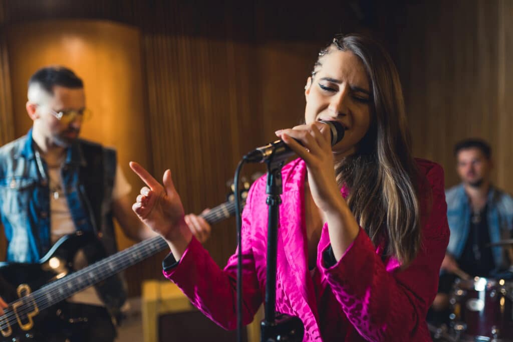 Eine Frau in einer leuchtend rosafarbenen Jacke singt leidenschaftlich in ein Mikrofon und liefert lebendige Musik, während zwei Musiker hinter ihr in einer schwach beleuchteten Halle Gitarre und Schlagzeug spielen.