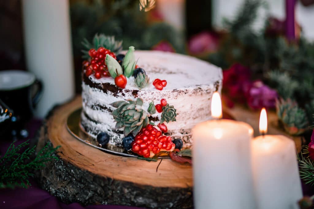 Eine rustikale, halbnackte, mit Beeren und Sukkulenten geschmückte Hochzeitstorte steht auf einer Holzplatte, drei weiße Kerzen brennen im Vordergrund und üppiges Grün im Hintergrund.