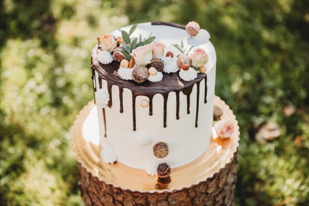 Eine weiß gefrostete Hochzeitstorte mit Schokoladentropfen, geschmückt mit rosa Rosen, Grünzeug, Baiser und verschiedenen Pralinen, steht auf einem goldenen Tablett auf einem Baumstumpf im Freien auf einer Wiese.