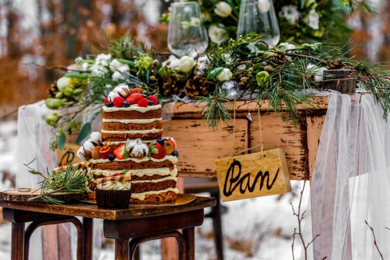 Eine rustikale Hochzeitstorte mit Sahne, Erdbeeren und Nüssen steht auf einem kleinen Holztisch. Dahinter steht ein dekorierter Holztisch mit Tannenzweigen, weißen Blumen, Tannenzapfen und einem hölzernen "Pan"-Schild vor einer verschneiten Außenkulisse.