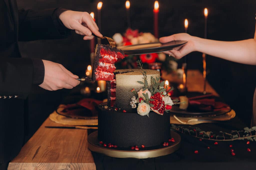 Zwei Personen schneiden und servieren ein Stück Hochzeitstorte aus rotem Samt mit dunklem, elegantem Zuckerguss und Blumendekoration, umgeben von Kerzen und festlichem Tischschmuck auf einem Holztisch.