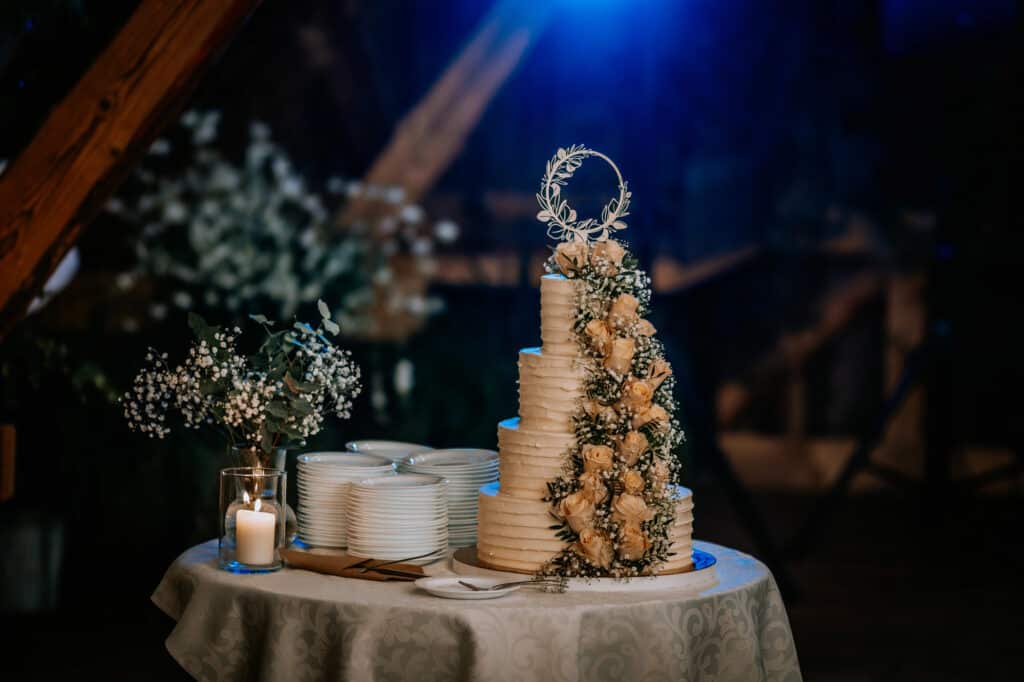 Ein runder Tisch mit einer hohen, gestuften Hochzeitstorte, geschmückt mit cremefarbenen Blumen, umgeben von gestapelten Tellern, Besteck, einer Kerze und einem Blumenarrangement, alles beleuchtet von dramatischem blauem Licht.