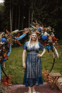 Stephanie Hansen, mit langen blonden Haaren, steht lächelnd in einem blauen Spitzenkleid vor einem Blumenbogen aus blauen, orangefarbenen und weißen Blüten, umgeben von grünem Gras und hohen Bäumen in dieser wunderschönen Umgebung.