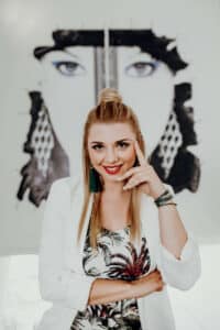 Eine lächelnde Frau mit langen blonden Haaren, die einen weißen Blazer und ein gemustertes Oberteil trägt, steht vor einem großen Schwarz-Weiß-Gemälde, das ein Gesicht zeigt. Stephanie Hansen stützt ihre Hand auf ihr Kinn und strahlt Selbstbewusstsein aus.