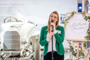Eine Frau in einem grünen Blazer spricht bei einer Indoor-Veranstaltung in ein Mikrofon. Hinter ihr bilden ein weißer Oldtimer und ein mit Grünpflanzen geschmücktes Schild mit der Aufschrift "Stephanie Hansen" eine einladende Kulisse.