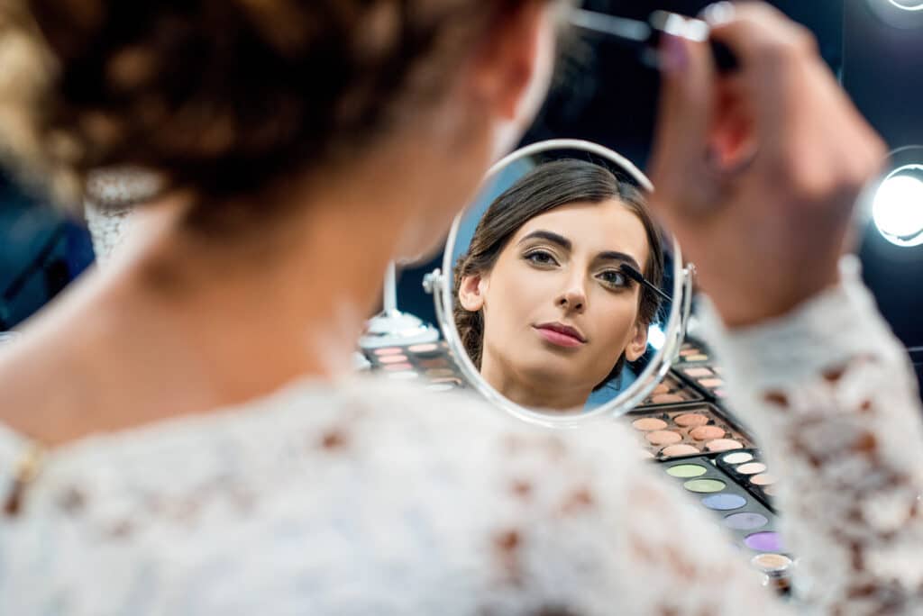 Eine Frau macht sich fertig, während sie Make-up aufträgt und dabei in einen kleinen runden Spiegel schaut, in dem ihr Spiegelbild deutlich zu sehen ist. Eine Lidschattenpalette und helle Lichter sind auf dem Tisch im Hintergrund zu sehen.