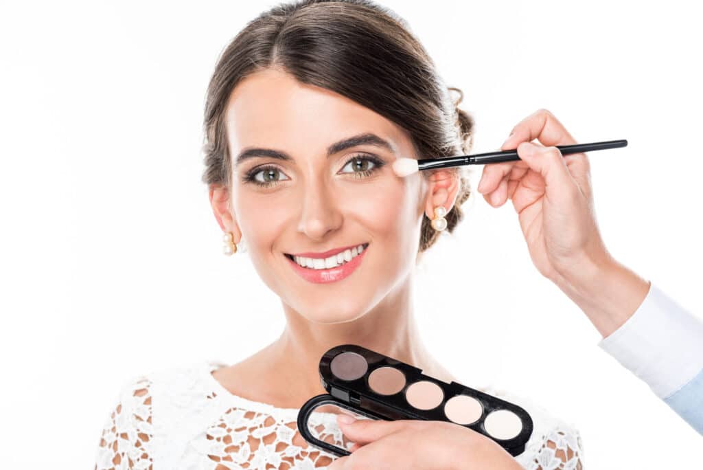 Eine Frau mit dunklem Haar in einer Hochsteckfrisur lächelt, während sie sich fertig macht und jemand mit einem Pinsel Lidschatten auf ihr Augenlid aufträgt. Sie trägt ein weißes Spitzenoberteil und Perlenohrringe, während sie eine Palette neutraler Lidschatten neben ihr Gesicht hält.