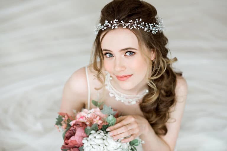 Eine Braut mit gewelltem Haar, die eine zarte Perlenkette und ein weißes Kleid trägt, hält einen Strauß rosa und weißer Blumen in der Hand, während sie sich fertig macht, nach oben schaut und sanft lächelt. Der Hintergrund ist sanft verschwommen.