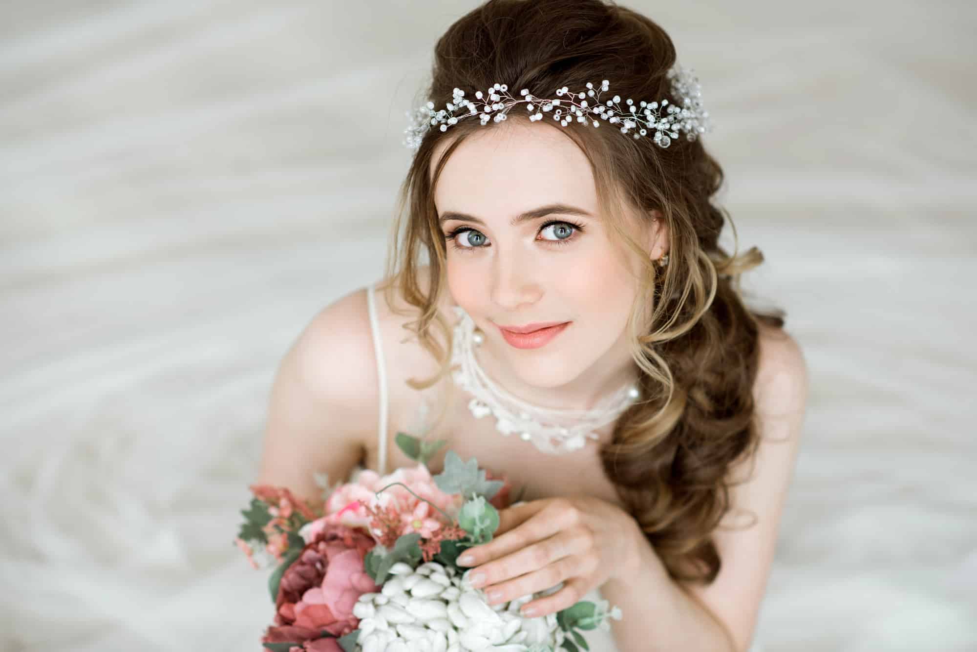 Eine Braut mit gewelltem Haar, die eine zarte Perlenkette und ein weißes Kleid trägt, hält einen Strauß rosa und weißer Blumen in der Hand, während sie sich fertig macht, nach oben schaut und sanft lächelt. Der Hintergrund ist sanft verschwommen.
