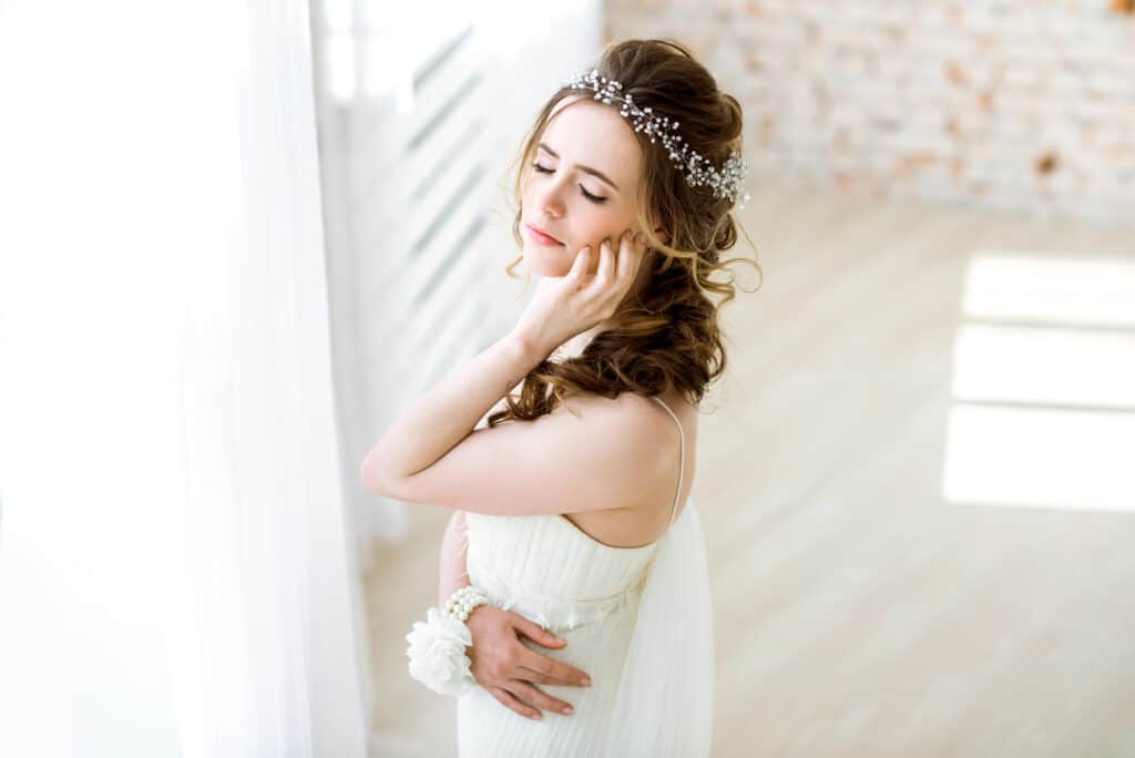 Eine Frau in einem weißen Kleid und mit Perlenkopfschmuck steht mit geschlossenen Augen da und berührt sanft ihr Gesicht, während sie in einem hellen, luftigen Raum einen ruhigen Getting Ready-Moment genießt und sich im sanften natürlichen Licht eines nahe gelegenen Fensters sonnt.