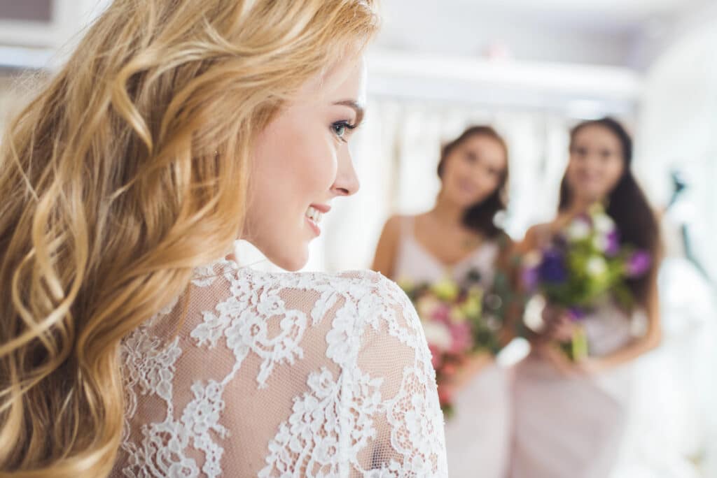 Eine Braut mit langen blonden Haaren und einem Hochzeitskleid aus Spitze lächelt während der Vorbereitungen und blickt auf zwei Brautjungfern in hellen Kleidern, die Blumensträuße im sanft beleuchteten Hintergrund halten.