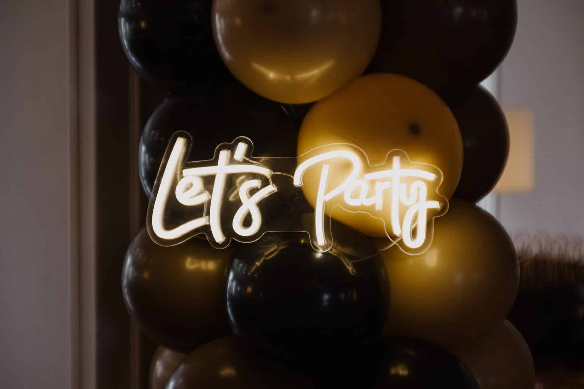 Eine Ansammlung schwarzer, goldener und gelber Luftballons bildet die Kulisse für Lebensfeiern. Davor leuchtet ein Neonschild mit der Aufschrift "Let's Party" in kursiven Buchstaben.