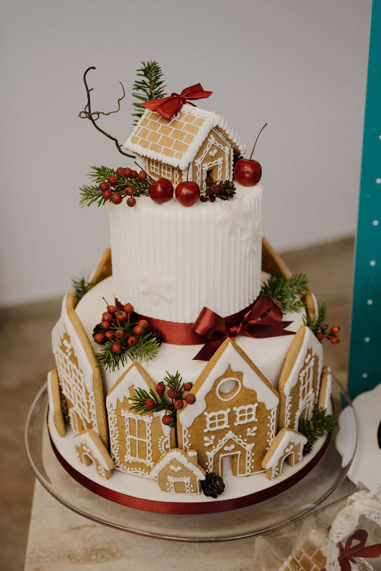 Eine festliche Torte für Lebensfeiern, verziert mit Lebkuchenhäusern, Tannenzweigen, roten Beeren und einer roten Schleife, gekrönt mit einem kleinen Lebkuchenhaus und Kirschen, steht auf einem gläsernen Kuchenständer.