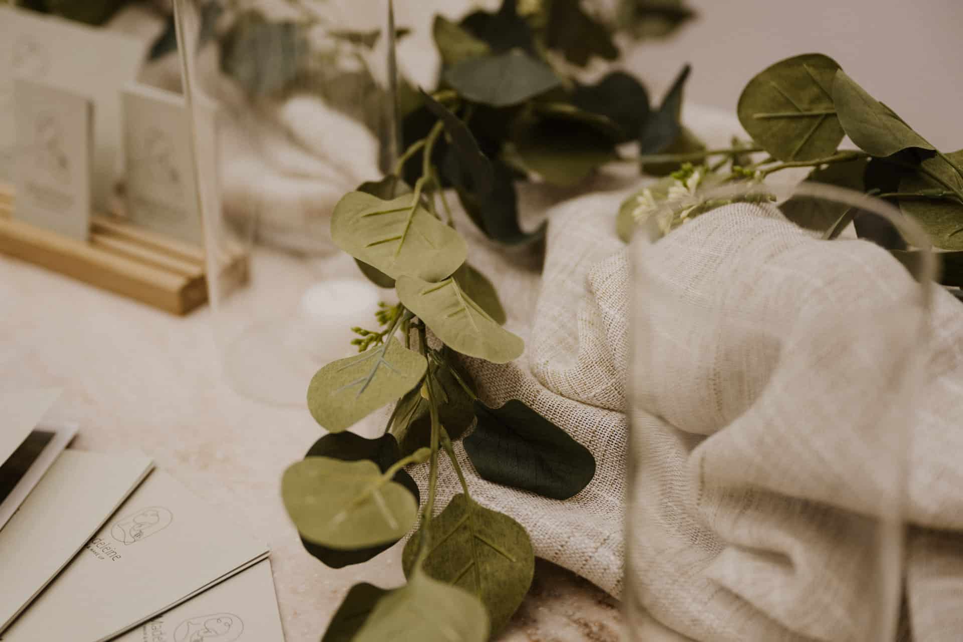 Eine Nahaufnahme von grünen Eukalyptusblättern auf einem beigen Leinentuch, mit Gläsern und Karten im Hintergrund, schafft eine weiche, natürliche und elegante Tischgestaltung - perfekt für intime Lebensfeiern.