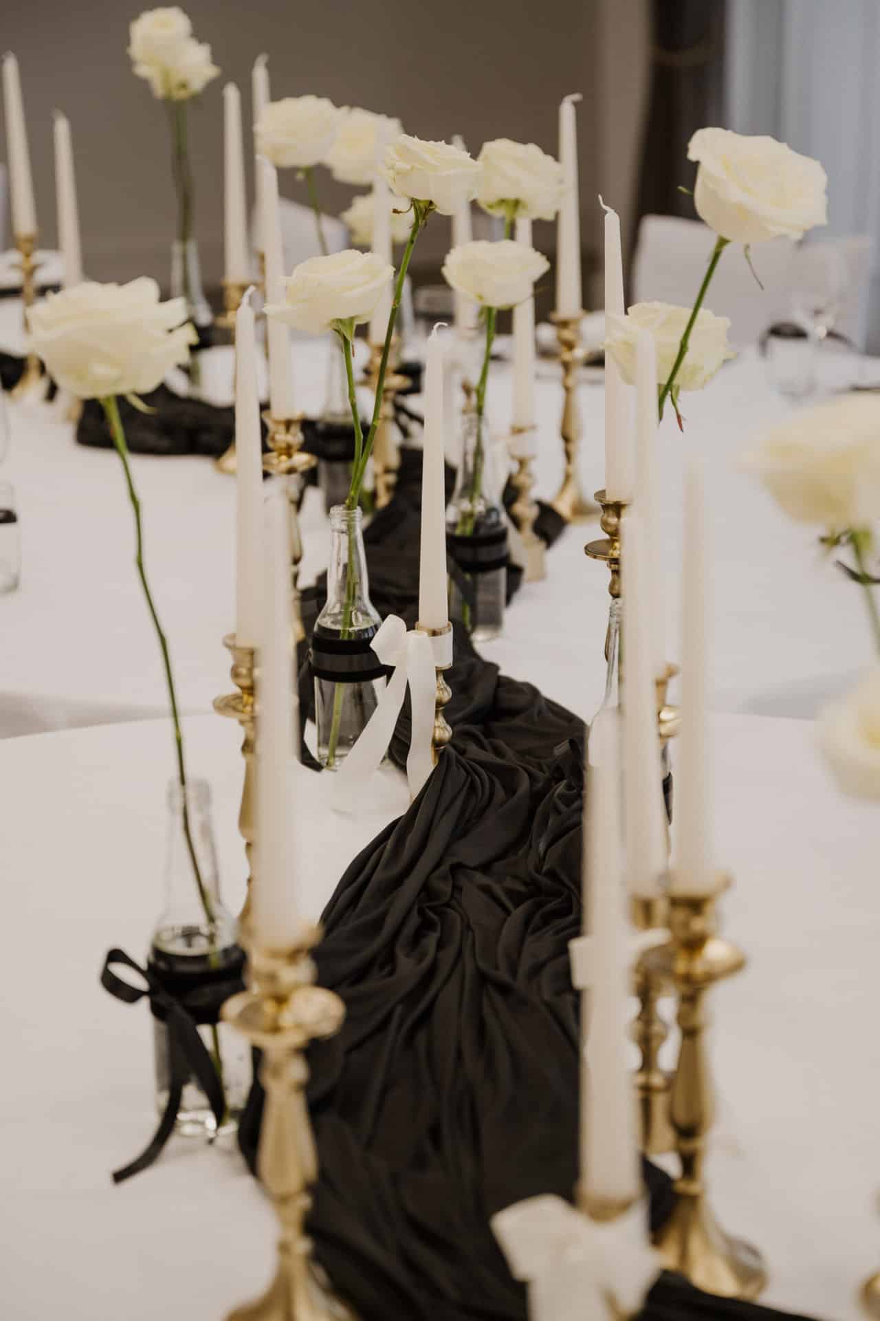 Ein für Lebensfeiern dekorierter Tisch mit goldenen Kerzenhaltern, weißen Spitzkerzen und Glasvasen mit einzelnen weißen Rosen. In der Mitte verläuft ein schwarzer Stoffläufer, der einen eleganten, monochromen Rahmen schafft.