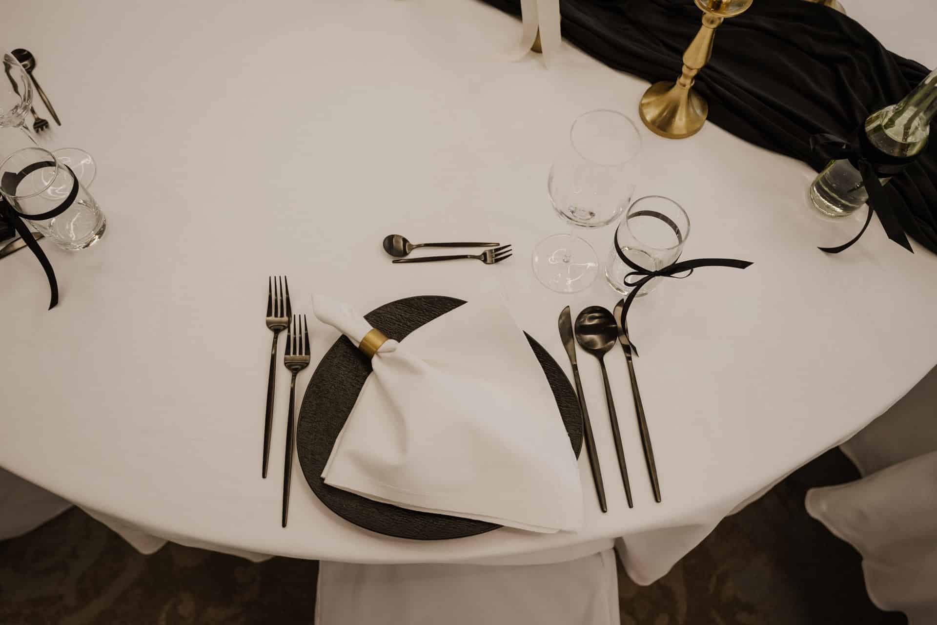 Ein formeller Tisch für Lebensfeiern besteht aus einem weißen Tischtuch, schwarzem Besteck, einem schwarzen Teller, einer weißen Serviette mit goldenem Ring, Glaswaren und einem schwarzen Tischläufer, der durch Kerzenhalter aus Messing und Glasflaschen akzentuiert wird.