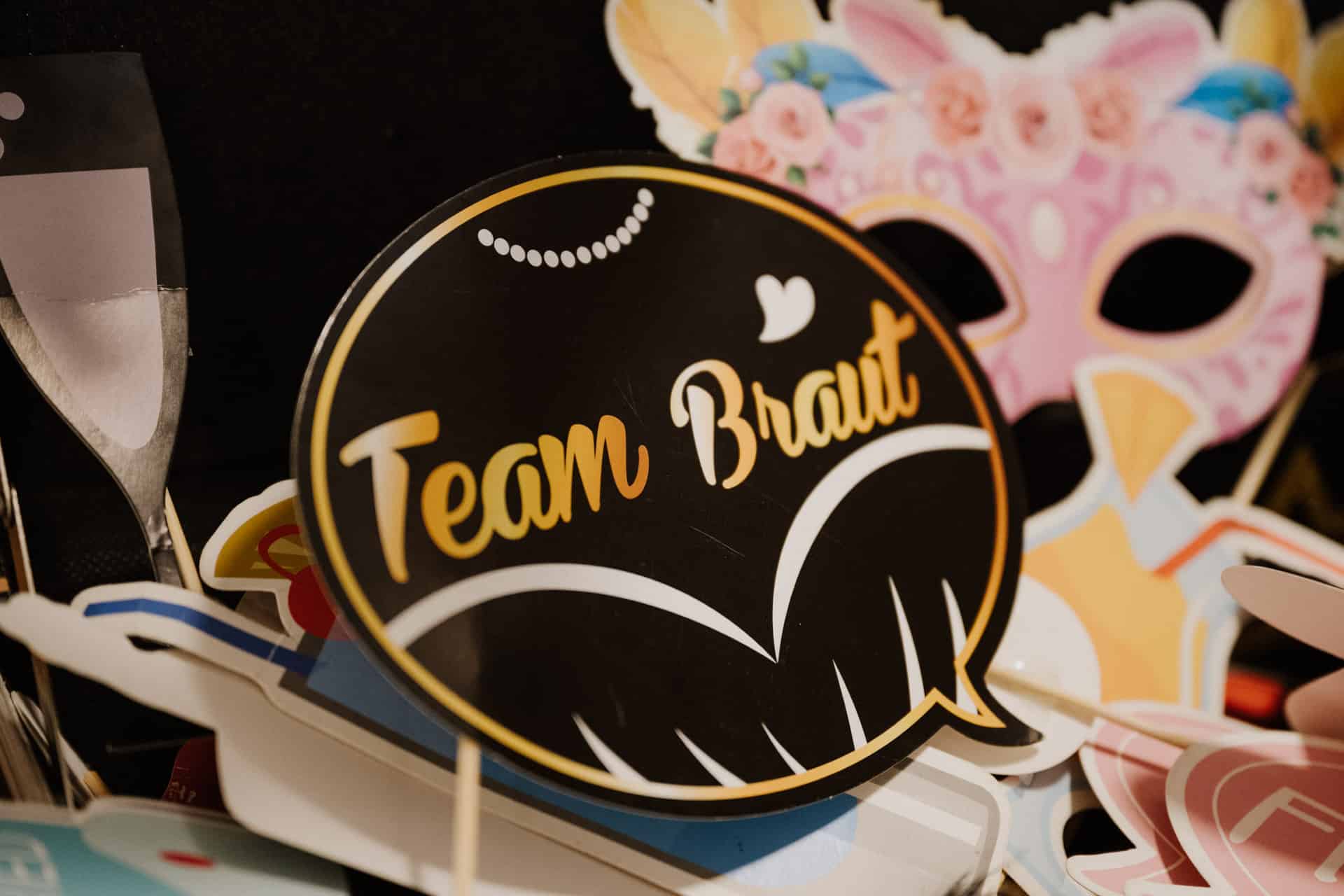 Eine Nahaufnahme eines dekorativen "Team Braut"-Schilds in goldenen und weißen Buchstaben, umgeben von bunten Papierrequisiten wie einer rosa Maske, einem Sektglas und Sprechblasen - perfekt für Lebensfeiern wie Hochzeiten oder Junggesellen-Fotokabinen.