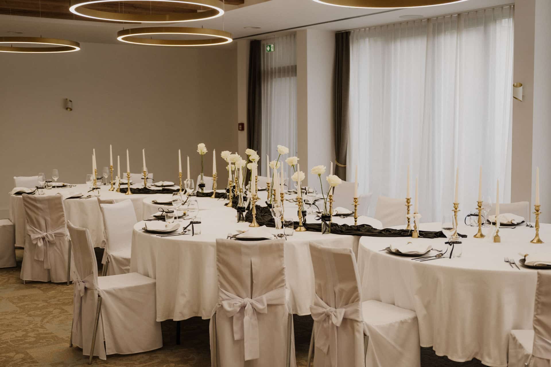 Elegante Banketttische in U-Form für Lebensfeiern, bedeckt mit weißen Tischtüchern und passenden Stuhlhussen, die mit Schleifen gebunden werden. Hohe Kerzenständer und weiße Blumen dienen als Mittelstücke, während große Fenster natürliches Licht hereinlassen.