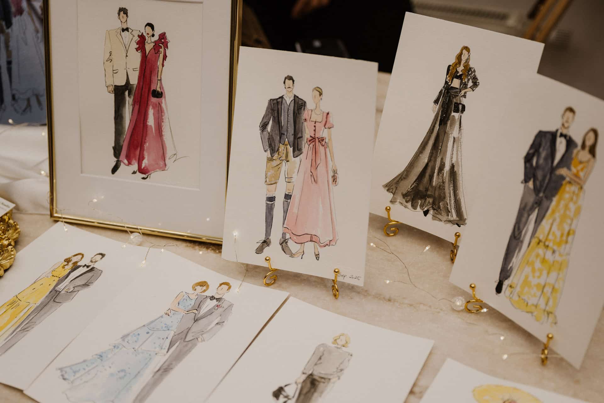 Eine Ausstellung mit Modeillustrationen, die elegant gekleidete Paare und Einzelpersonen in förmlicher Kleidung, einschließlich Kleidern und Anzügen, auf einem Tisch mit goldenen Ständern und weichen Lichterketten zeigt - perfekt für Lebensfeiern.