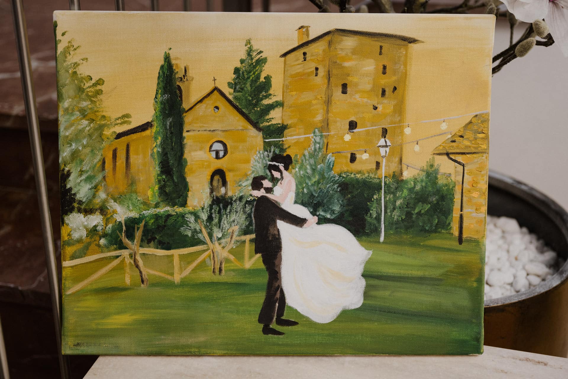Eine gemalte Leinwand zeigt ein Brautpaar, das während seiner Lebensfeiern im Freien auf einer Wiese tanzt. Im Hintergrund sind gelbe Gebäude, grüne Bäume, ein Holzzaun und Lichterketten zu sehen. Die Szene wirkt lebendig und romantisch.