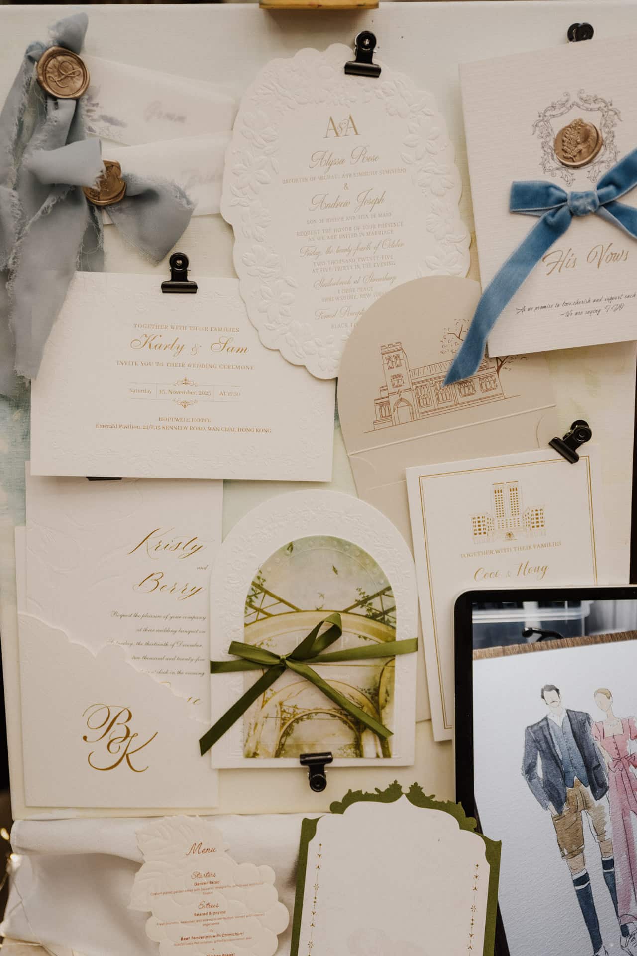 Ein Display mit eleganten Hochzeitseinladungen und Karten für Lebensfeiern, angeordnet auf einem Brett mit Klammern. Die Karten sind mit geprägten Motiven, Goldschrift und Zierbändern in Blau, Grün und Beige versehen. Eine Modeillustration ist teilweise sichtbar.