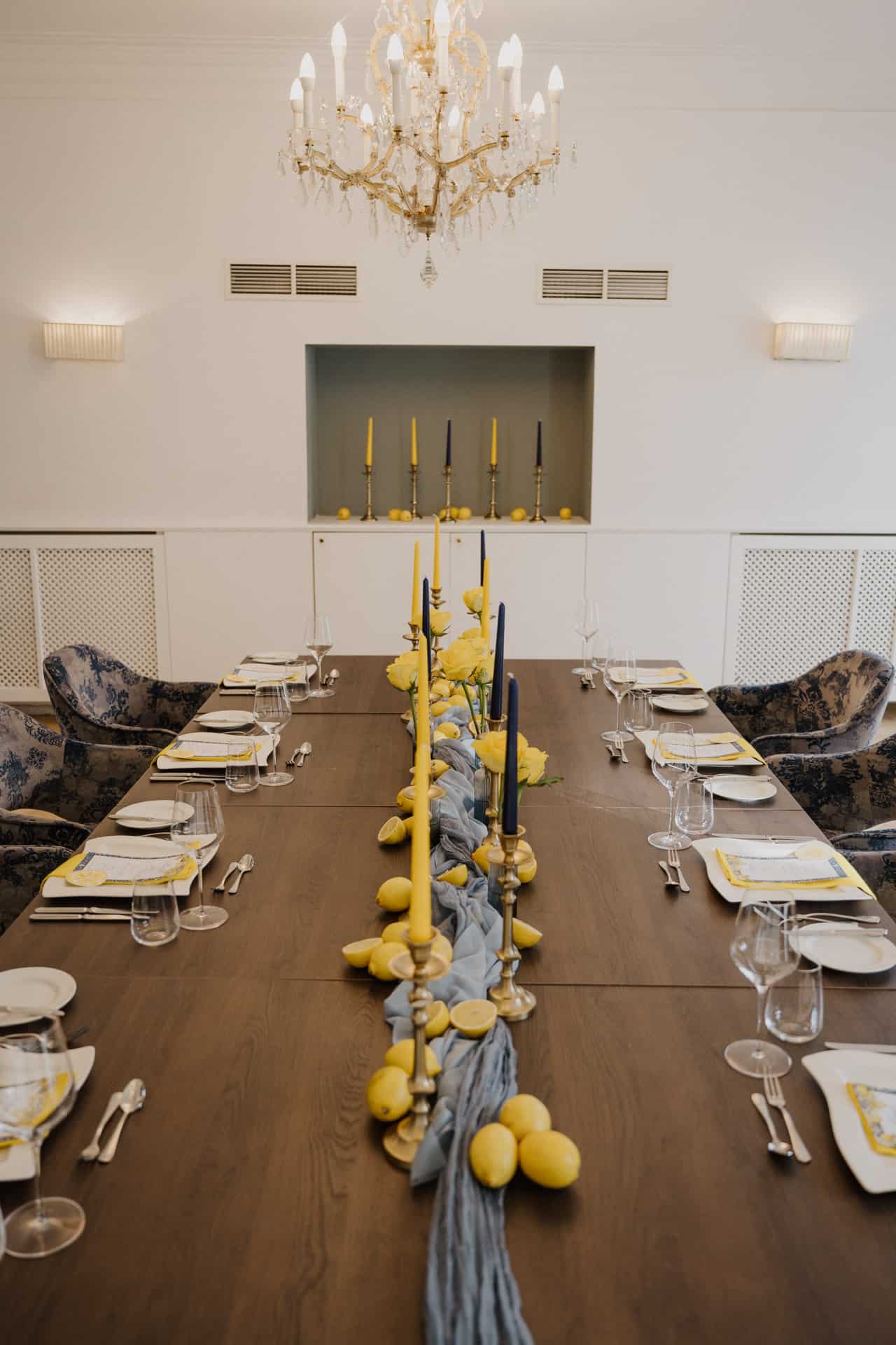 Ein langer Esstisch ist elegant für Lebensfeiern gedeckt, mit gelben und blauen Kerzen, Zitronen und blauem Stoff als Mittelstück. Teller, Gläser und Besteck sind fein säuberlich unter einem Kronleuchter angeordnet, während im Hintergrund Kerzen auf einem Regal stehen.