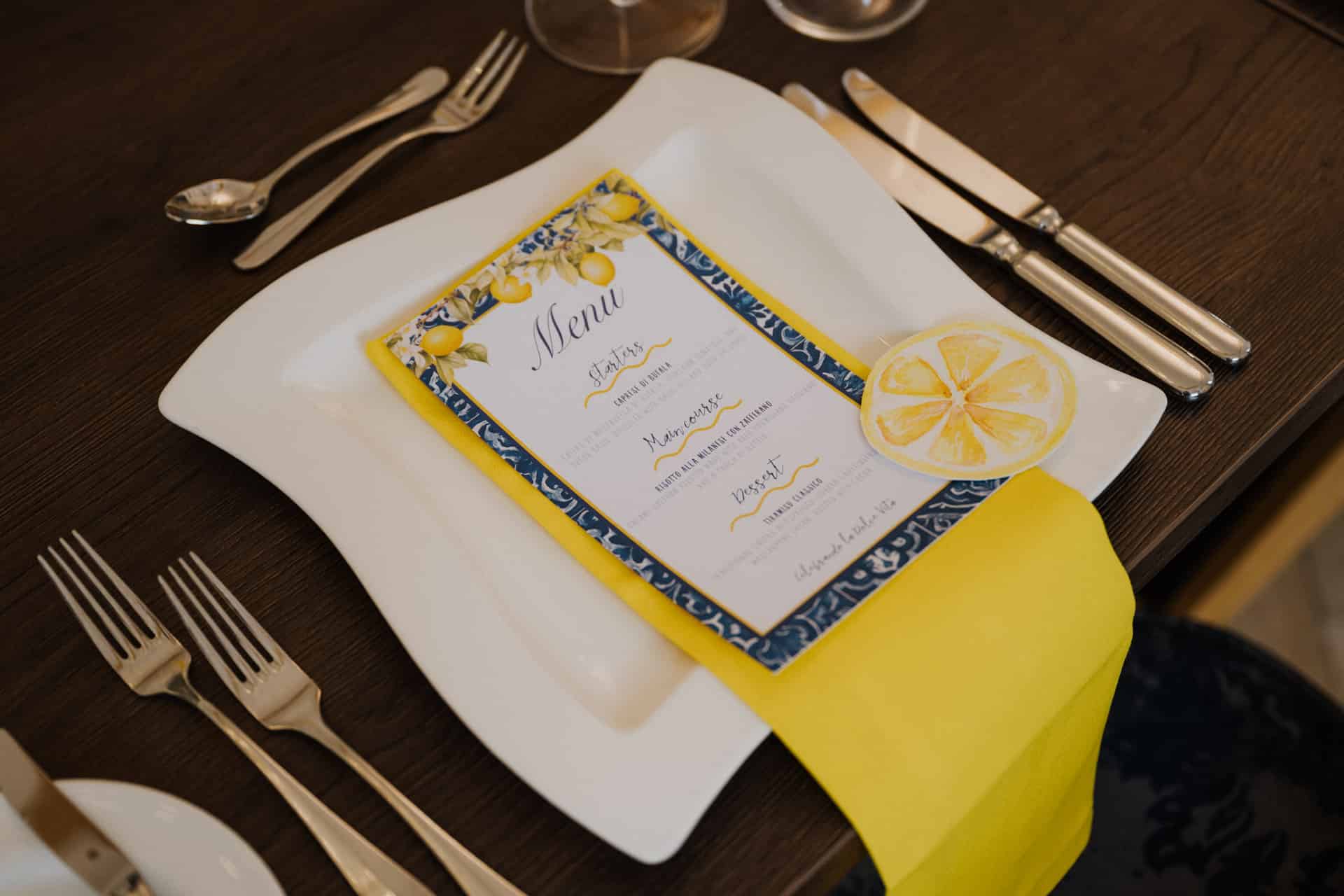 Ein formeller Tisch für Lebensfeiern besteht aus einem weißen Teller mit einer gelben Serviette, einer gedruckten Speisekarte mit blauen und gelben Akzenten und einer Zitronenscheibe als Dekoration. Silbernes Besteck und Weingläser sind auf einem Holztisch angeordnet.