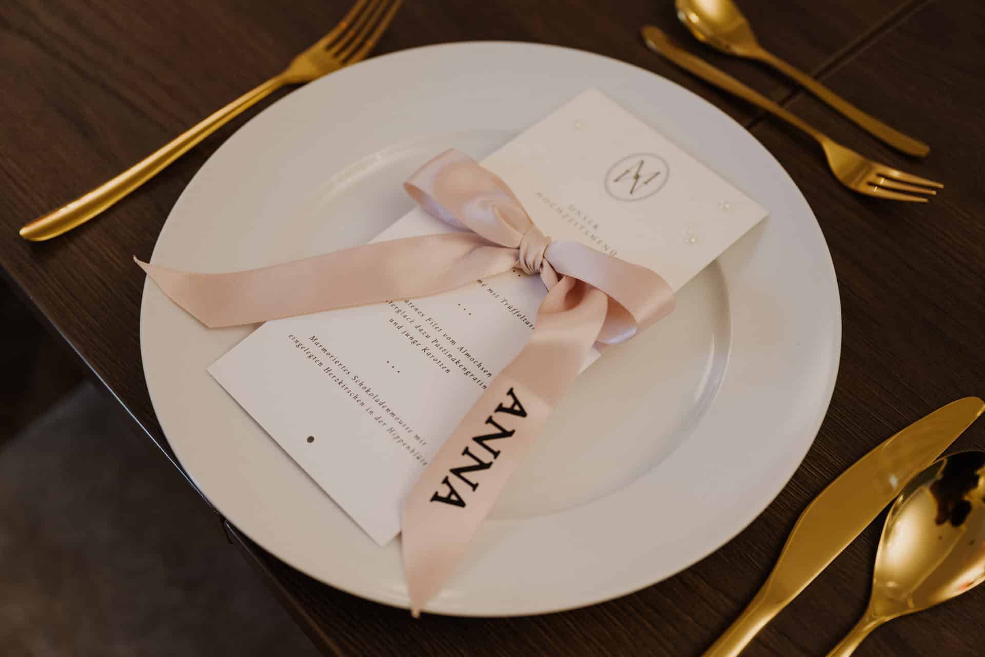 Auf einem weißen Teller auf einem Holztisch liegt eine Menükarte mit einem zartrosa Band, das mit einer Schleife gebunden und mit "Anna" bedruckt ist. Goldene Utensilien sind ordentlich um den Teller herum angeordnet, perfekt für elegante Lebensfeiern.