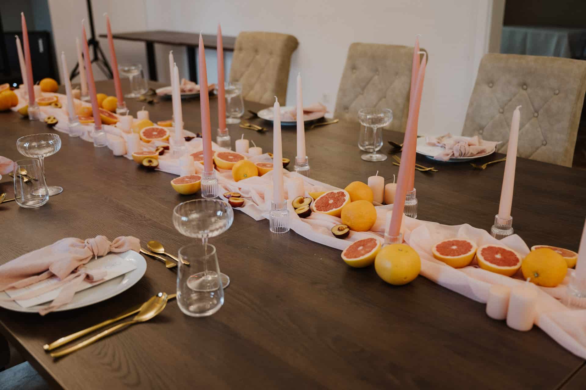 Ein Esstisch ist mit goldenem Besteck, Gläsern und neutralen Servietten gedeckt. Perfekt für Lebensfeiern ist der Tafelaufsatz mit rosafarbenen Kerzen, Stoff und halbierten Grapefruits und Pflaumen für einen sanften, eleganten und frischen Look.