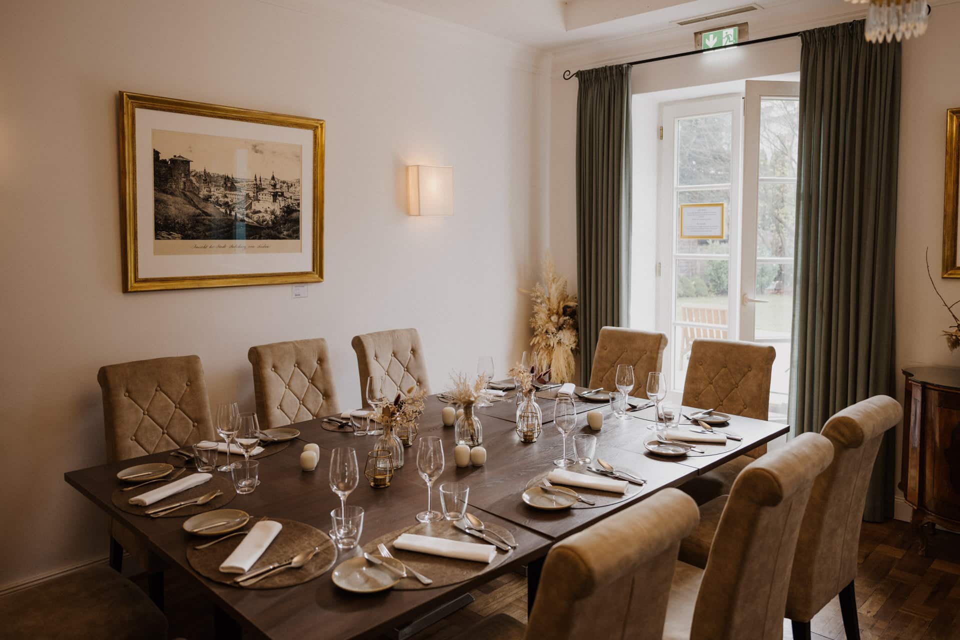 Ein Esszimmer mit einem großen Holztisch für zehn Personen, beigefarbenen Polsterstühlen, ordentlich angeordnetem Geschirr und Gläsern - ein einladender Rahmen für Lebensfeiern. An der Wand hängt ein Gemälde, und Glastüren führen nach draußen in einen Garten.
