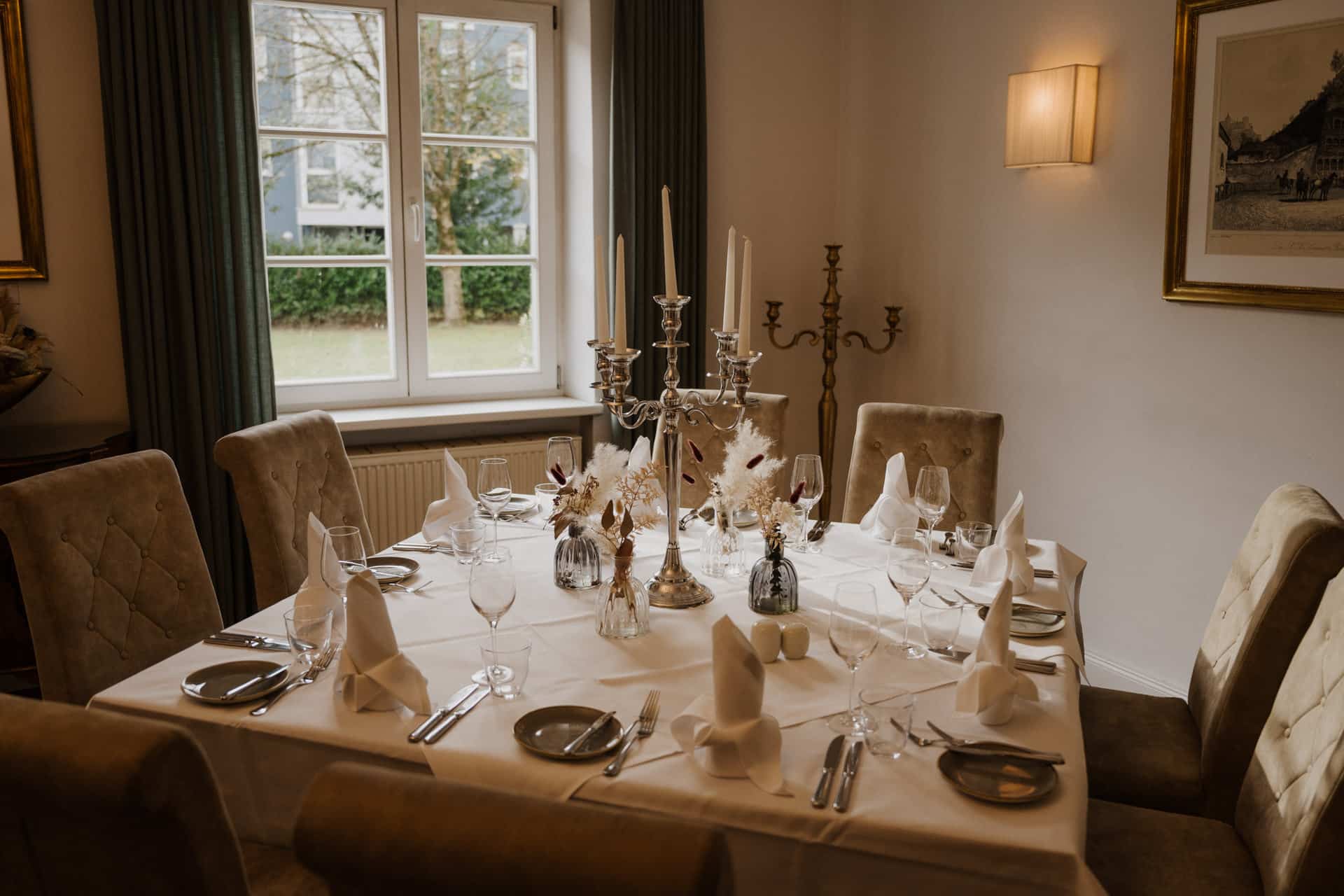 Ein formell gedeckter Esstisch mit gefalteten weißen Servietten, Gläsern, Tellern und Silberbesteck ist perfekt für Lebensfeiern. Elegante Kandelaber und weiße Blumenarrangements schmücken die Mitte; sechs gepolsterte Stühle umgeben den Tisch in einem hellen Raum.