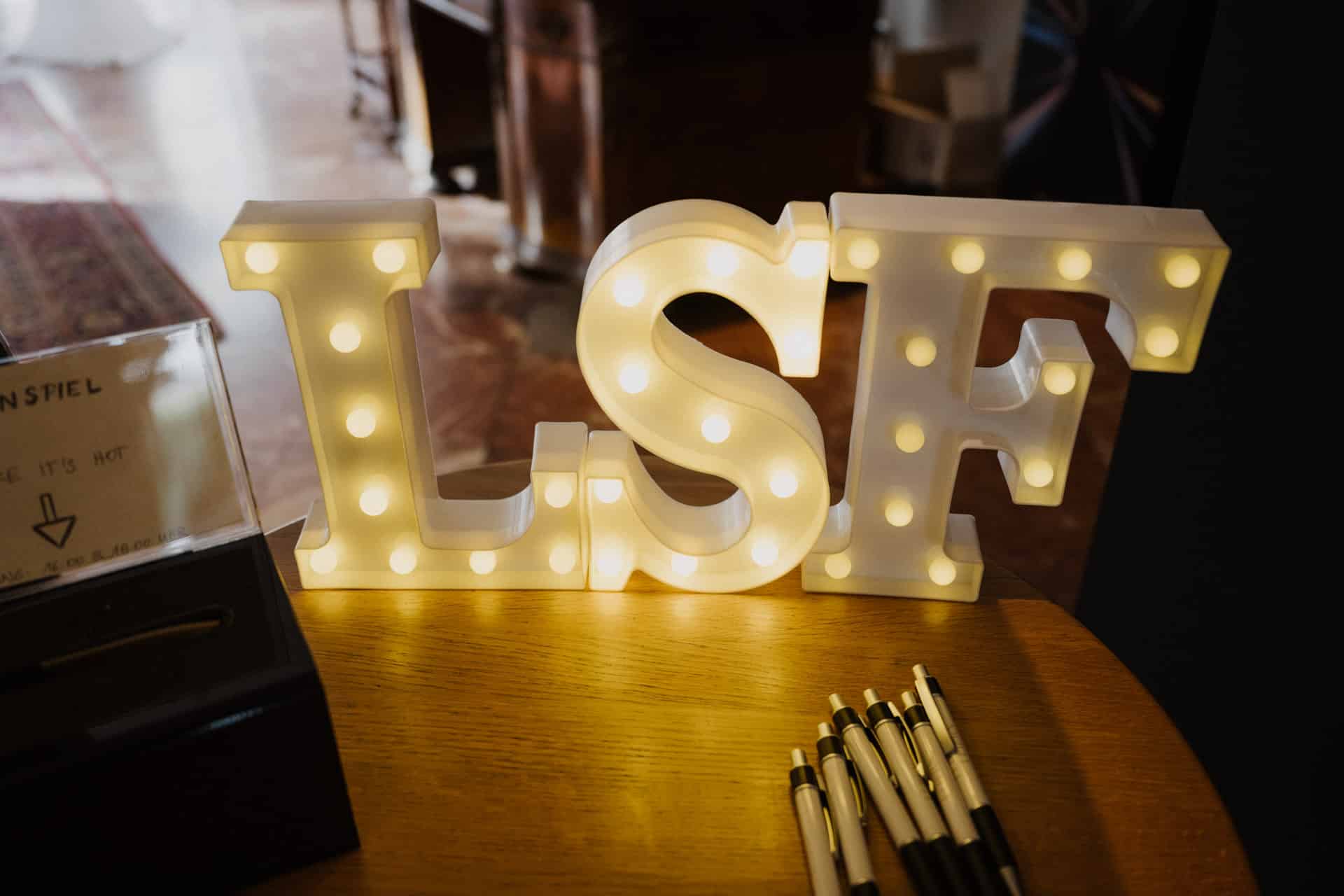 Große weiße Buchstaben mit der Aufschrift "LSF" - kurz für Lebensfeiern - leuchten mit kleinen Glühbirnen auf einem Holztisch. In der Nähe sind im Vordergrund ein Kasten mit Stiften und ein teilweise sichtbares Schild zu sehen.