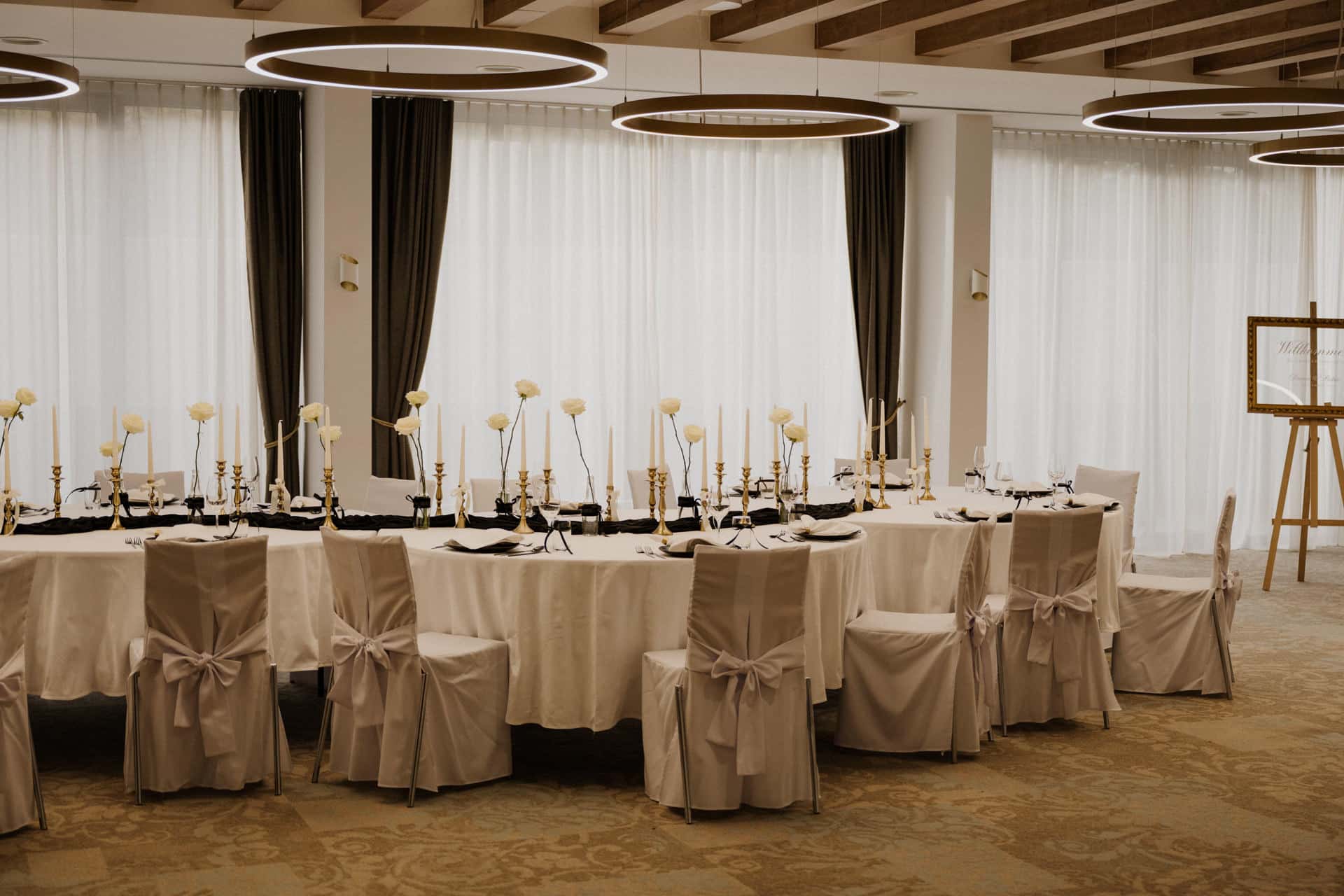 Eine lange Banketttafel mit weißer Tischdecke, elegante weiße Stühle, die mit Bändern gebunden sind, goldene Kerzenständer und weiße Blumen bilden in einem hellen Raum mit großen Fenstern und kreisrunden Deckenleuchten einen prächtigen Rahmen für Lebensfeiern.