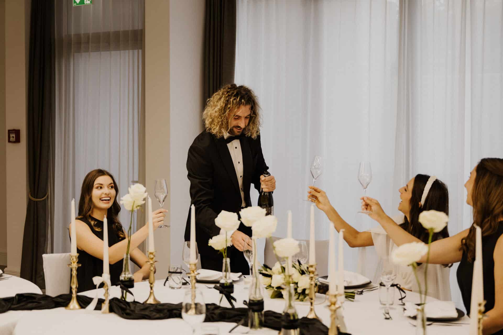 Ein Mann im Smoking öffnet eine Champagnerflasche an einem eleganten Tisch, der für Lebensfeiern gedeckt ist, während drei Frauen die Gläser zum Anstoßen hochhalten. Weiße Blumen, Kerzen und schwarze Tischläufer sorgen für ein festliches Ambiente.